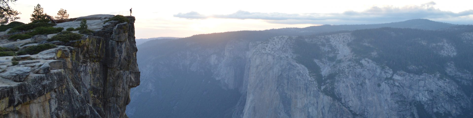 Image: wide view of a person standing on a cliff face looking out at a valley surrounded by mountains