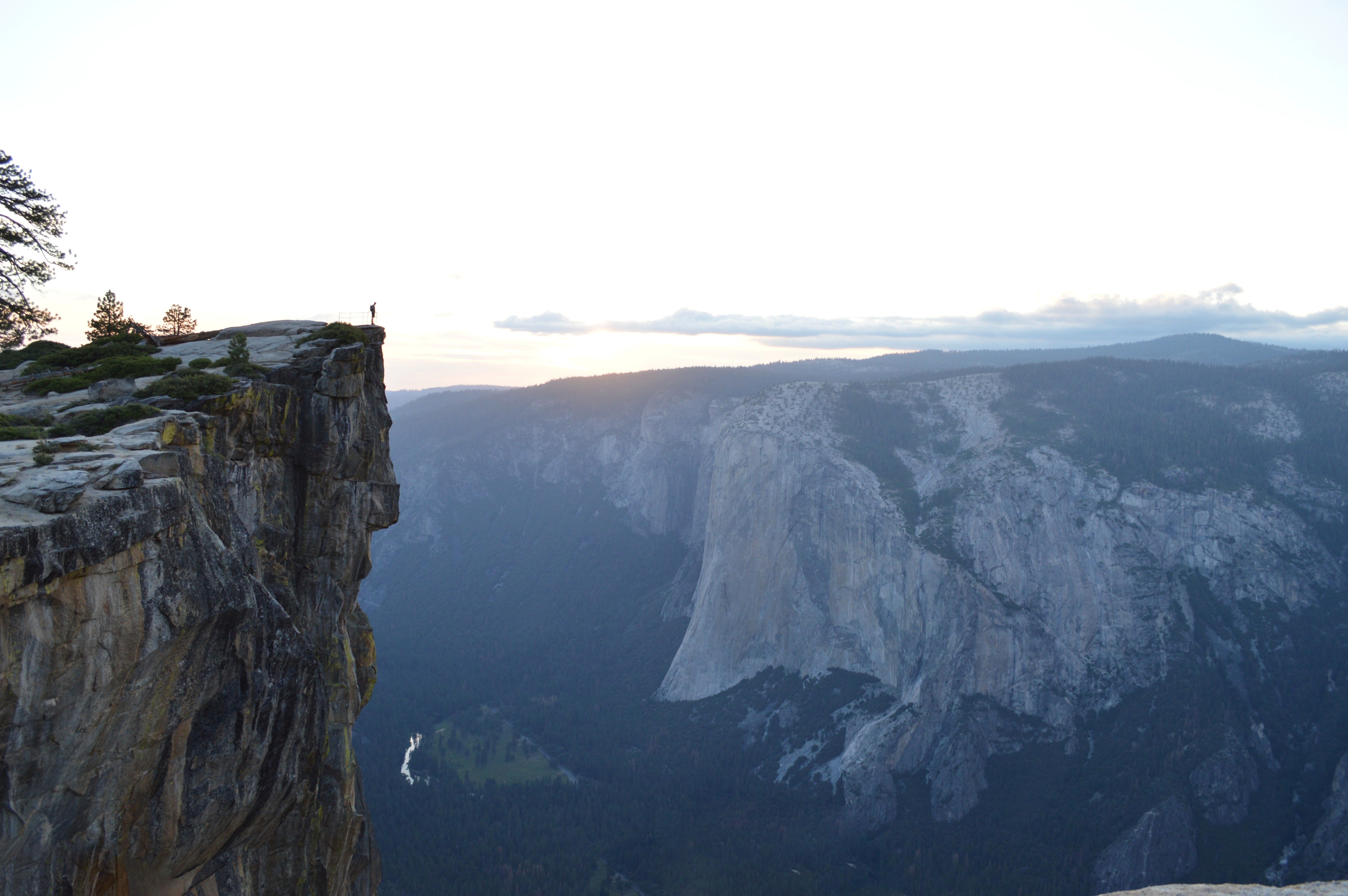 Image: wide view of a person standing on a cliff face looking out at a valley surrounded by mountains